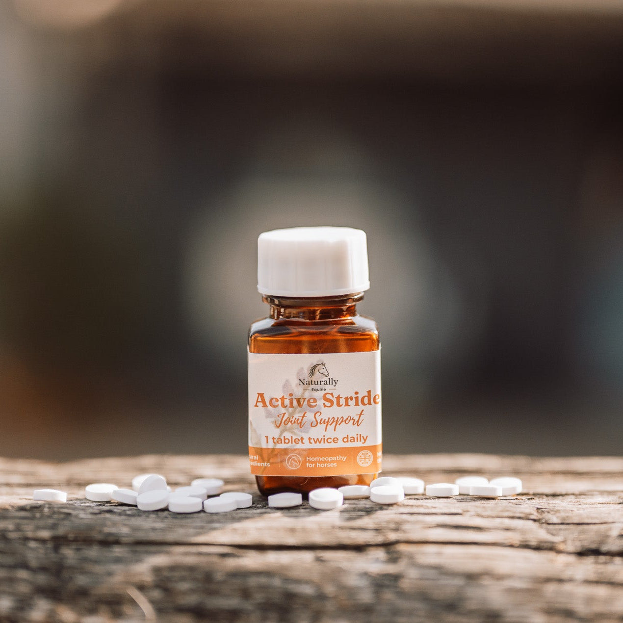 A small amber glass bottle with a white cap labeled 'Active Stride Joint Support' sits on a weathered wooden surface with scattered white tablets around it, against a softly blurred outdoor background.