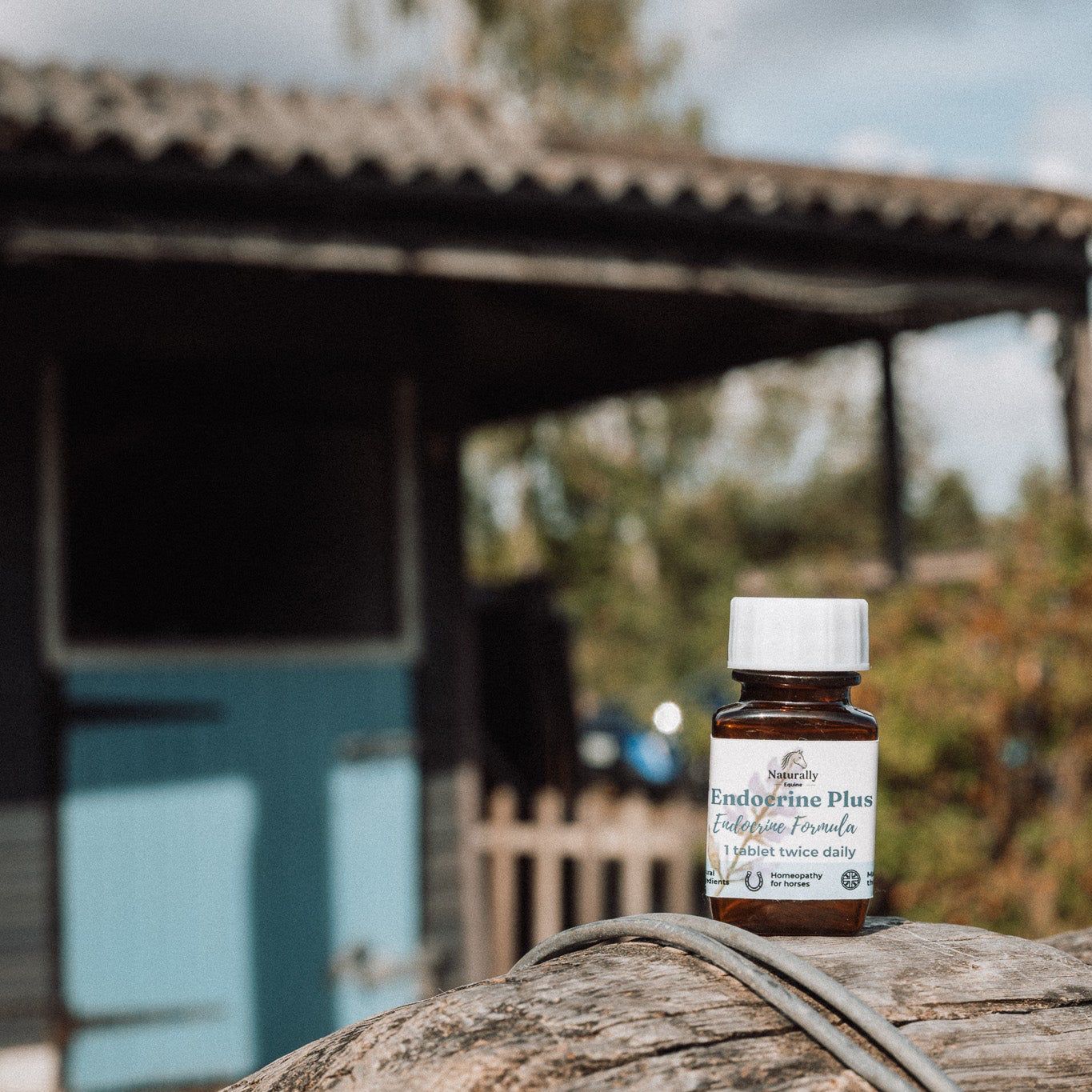 A small amber glass bottle with a white cap  sits on a weathered wooden barrel outdoors, with a stable and cloudy sky blurred in the background.