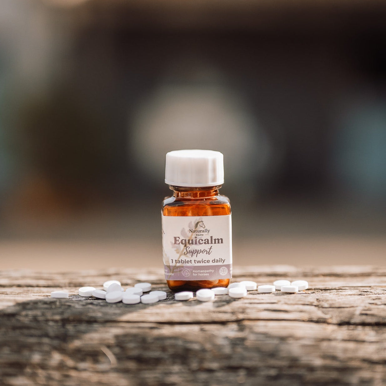 A small amber glass bottle labeled 'Equicalm Support' with a white cap, standing on a weathered wooden surface with scattered white tablets around it. The background is softly blurred.