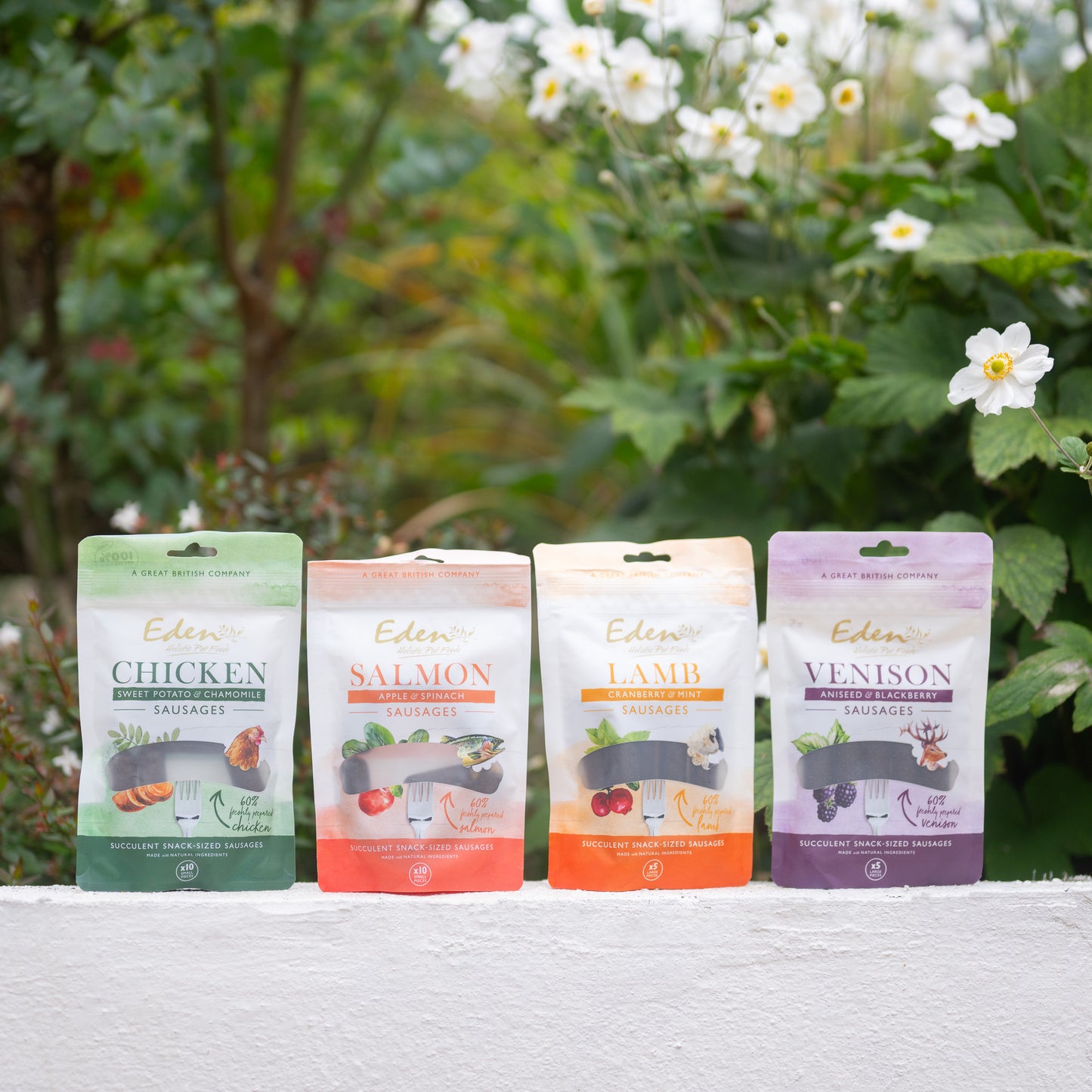 Eden Sausage Treats: chicken, salmon, lamb, and venison sausages in colorful packaging near flowers.