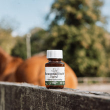 A small amber glass bottle of  Seasonal Itch Support sits on a weathered wooden fence with a blurred horse and outdoor trees in the background.