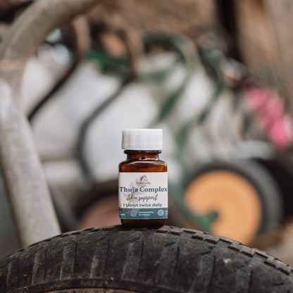 A small amber glass bottle with a white cap and a white label reading 'Thuja Complex' stands on top of a black tire in a rustic outdoor setting with blurred metal and wood in the background.