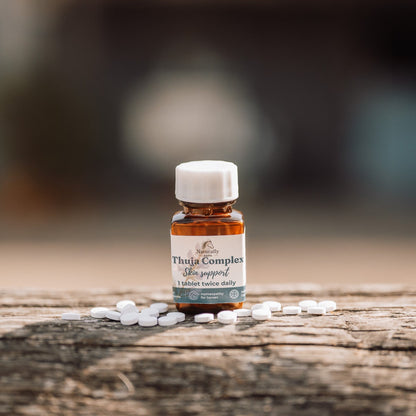 A small amber glass bottle labeled 'Thuja Complex' with a white cap, standing on a weathered wooden surface with scattered white tablets around it. The background is heavily blurred, showing an outdoor bokeh.