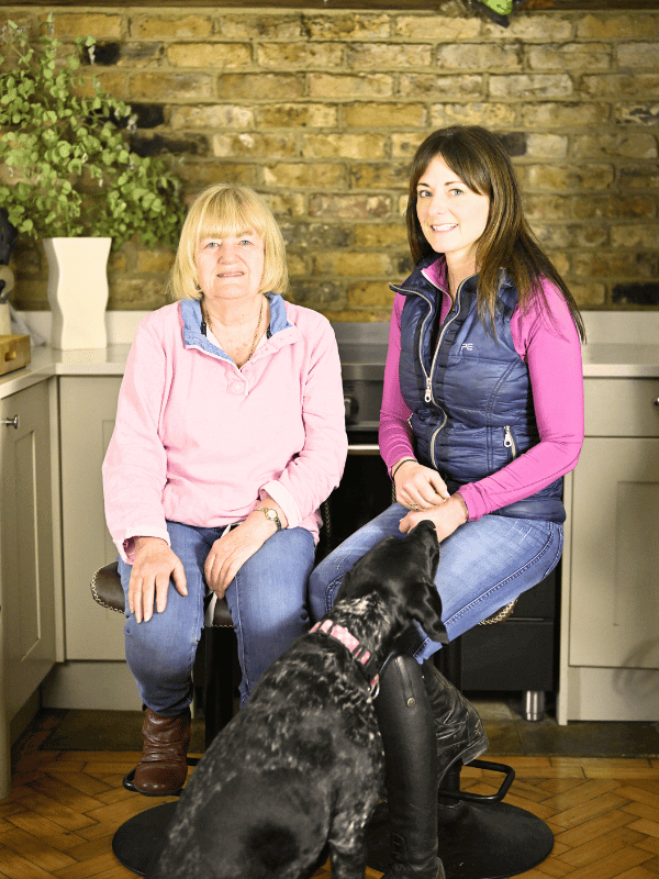 Two women sitting on stools in a kitchen with a dog in front, showcasing a cozy home environment.